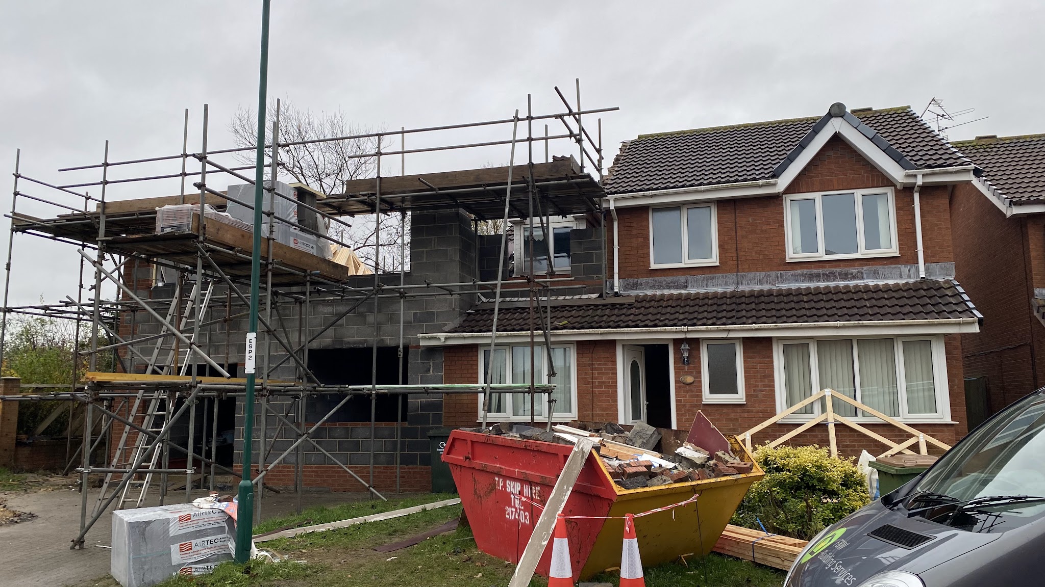 Blockwork walls of a two-storey side extension going up under scaffolding, with a skip and an F&W Building Services van in front